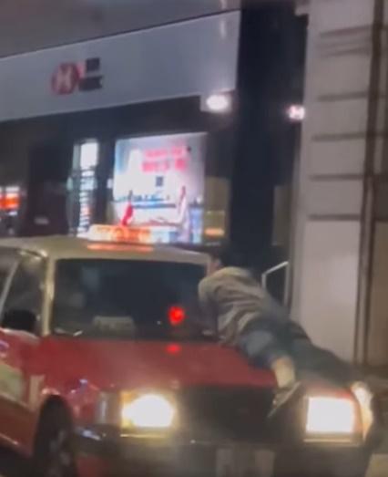 一名男子俯伏在一輛市區的士車頭上大叫:「你夾住我隻手呀!」並呼喝「大晒呀!」。 (的士司機資訊網 Taxi@FB)
