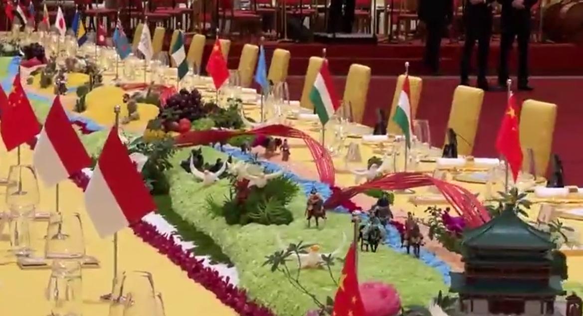 逾40米長桌上每個位置都擺放了不同國家的國旗,枱中央設計為「一帶一路」標誌,一側以是古代絲綢之路景象。(微博)