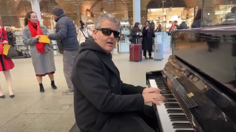 英國鋼琴家「K博士」卡瓦納(Brendan Kavanagh)上周五(19日)在倫敦St. Pancras火車站的街頭鋼琴進行公眾表演,期間身後不遠處站著數名手持中國國旗的遊客