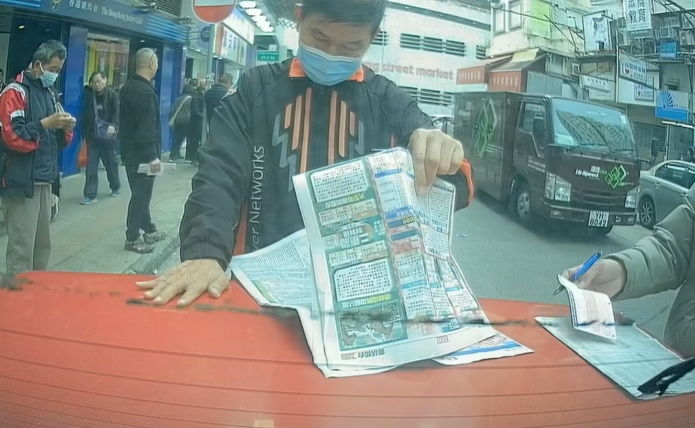 兩名中年馬迷於路邊手持原子筆,以車尾當書枱「刨馬經」,身穿杏色外套的男子更以車尾冚做書枱劃飛,未有留意司機已返回車上。(facebook群組「馬路的事討論區」影片截圖)