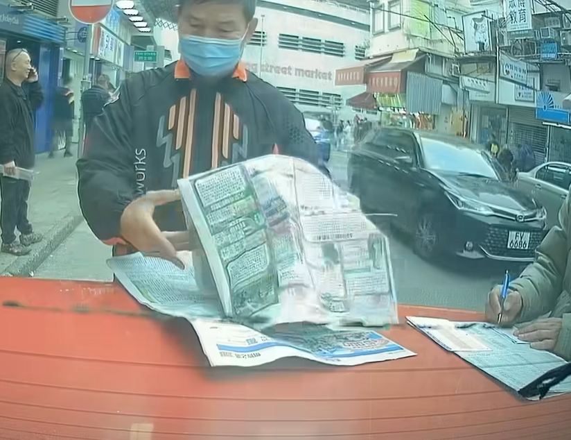 兩名中年馬迷於路邊手持原子筆,以車尾當書枱「刨馬經」,身穿杏色外套的男子更以車尾冚做書枱劃飛,未有留意司機已返回車上。(facebook群組「馬路的事討論區」影片截圖)