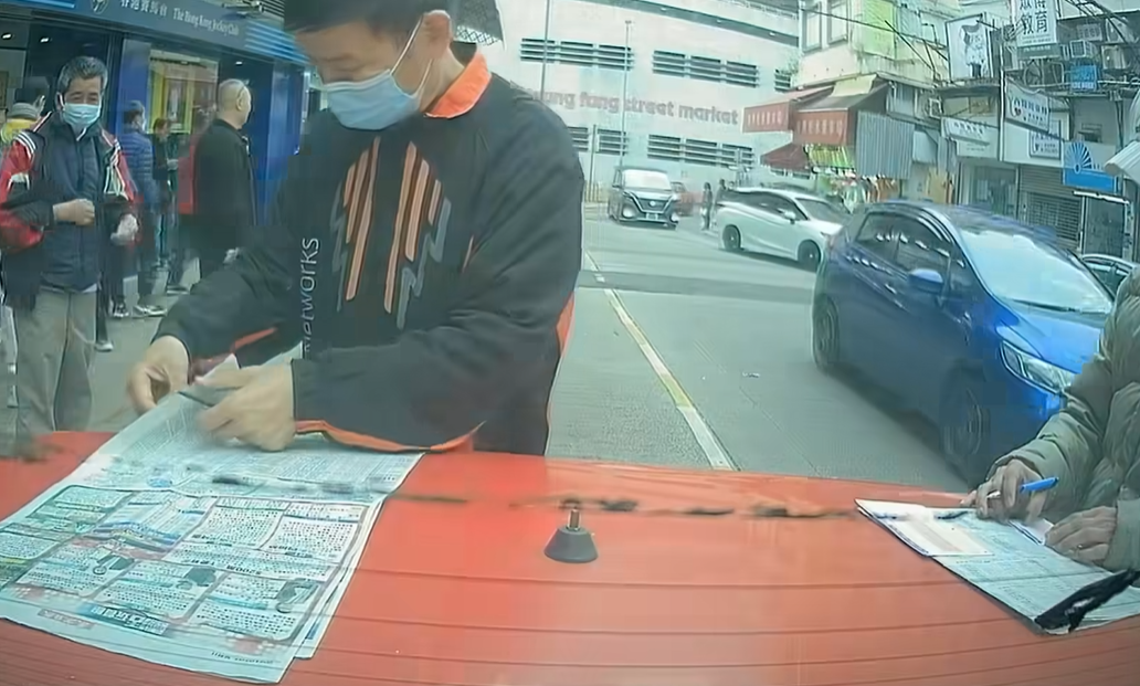 兩名中年馬迷於路邊手持原子筆,以車尾當書枱「刨馬經」,身穿杏色外套的男子更以車尾冚做書枱劃飛,未有留意司機已返回車上。(facebook群組「馬路的事討論區」影片截圖)