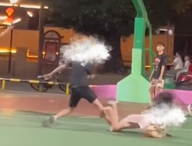 一名穿黑衣短褲的少年於籃球場急步助跑數步,飛身朝前方步行的一名穿粉色衫少女飛踢。(影片截圖)