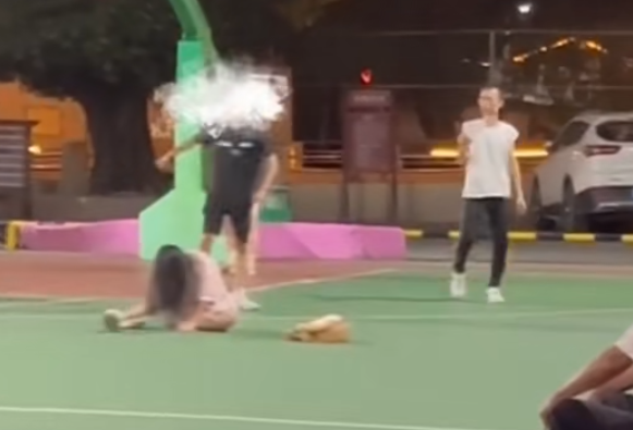 一名穿黑衣短褲的少年於籃球場急步助跑數步,飛身朝前方步行的一名穿粉色衫少女飛踢。(影片截圖)