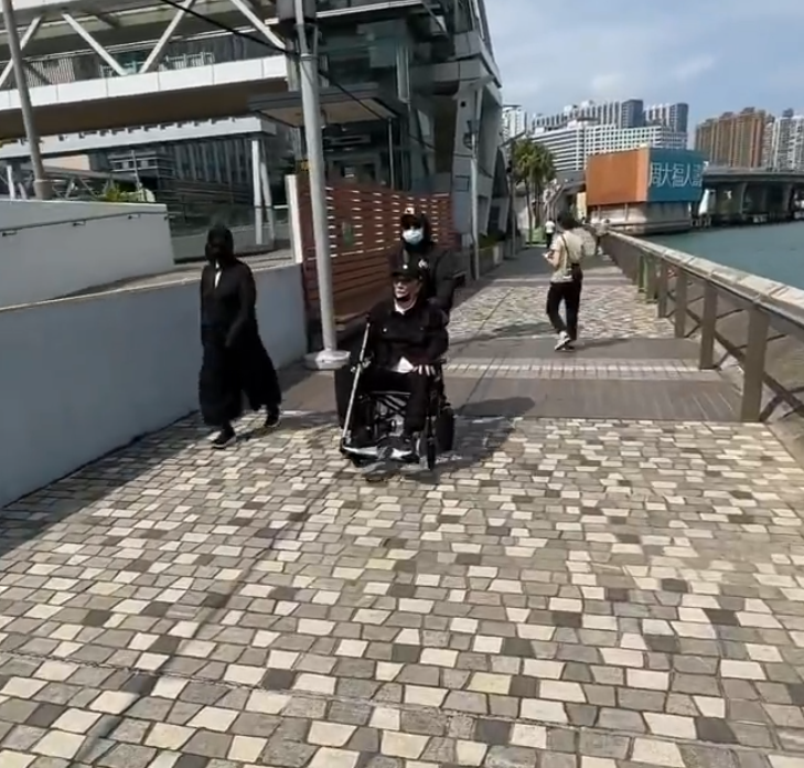 片中的謝賢戴上黑色Cap帽、黑超,又以全黑造型坐在輪椅上於尖沙咀散步,身邊有兩位同樣以全黑打扮的人在身旁陪伴及照顧他(影片截圖)