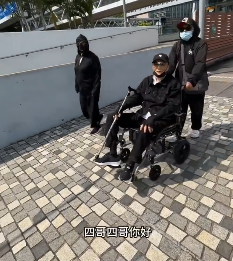 片中的謝賢戴上黑色Cap帽、黑超,又以全黑造型坐在輪椅上於尖沙咀散步,身邊有兩位同樣以全黑打扮的人在身旁陪伴及照顧他(影片截圖)