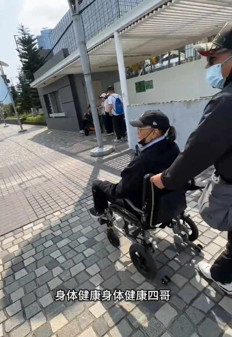片中的謝賢戴上黑色Cap帽、黑超,又以全黑造型坐在輪椅上於尖沙咀散步,身邊有兩位同樣以全黑打扮的人在身旁陪伴及照顧他(影片截圖)