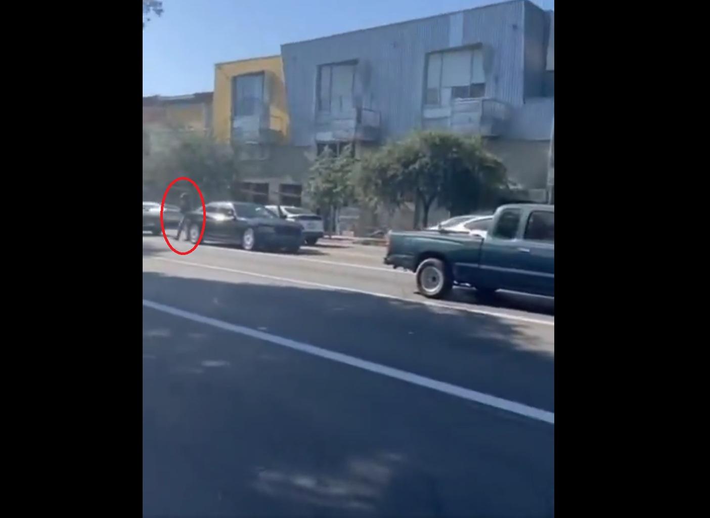 本月18日在加州帕薩迪納(Pasadena),一名疑似ICE男探員(紅圈)下車,拿槍指嚇在馬路另一邊拍攝的民眾。(互聯網)