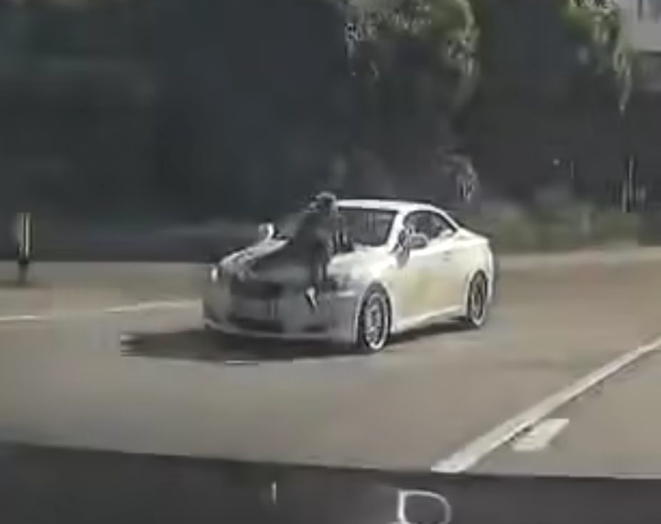 片段中一輛白色私家車沿粉錦公路行駛,一名穿黑衣的人伏在該車頭上(影片截圖)