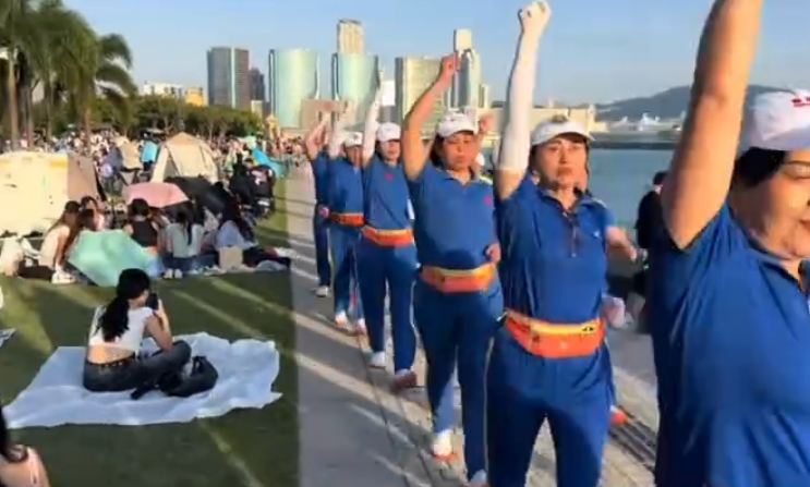 內地屢引發爭議的「暴走團」殺入香港,現身西九文化區徒步和做伸展運動,沿途大聲播放音樂影片,惹來旁邊市民及遊客側目。(影片截圖)